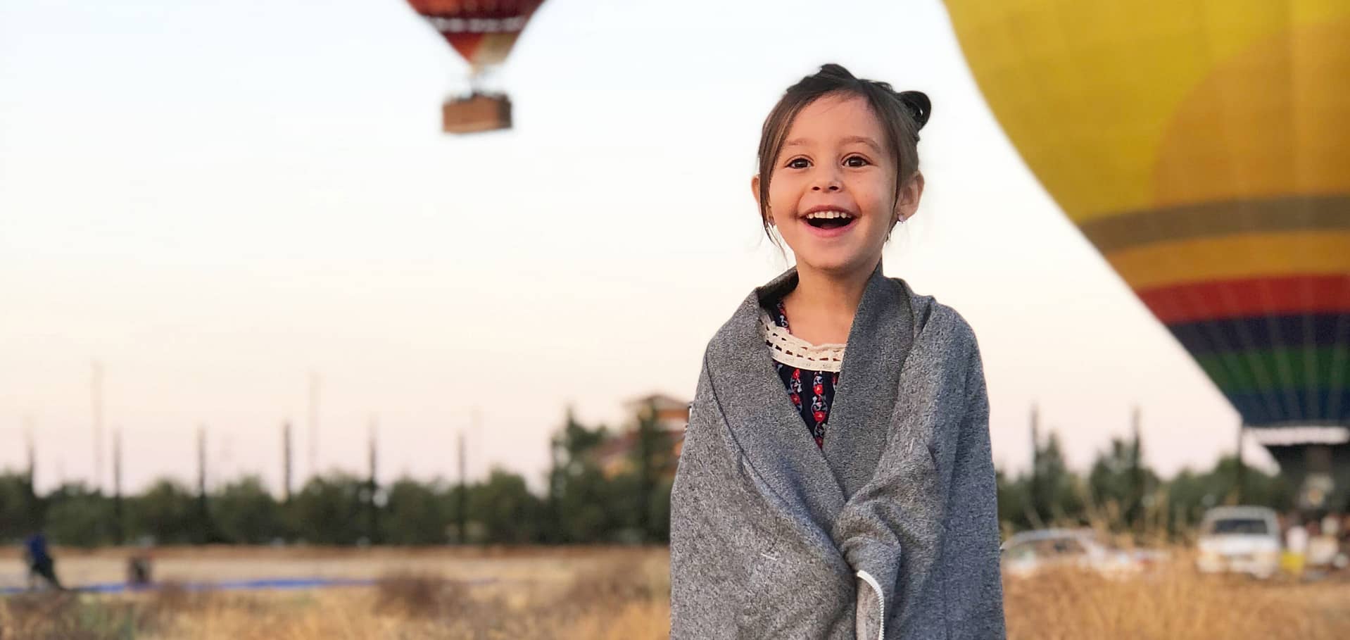 Happy girl smiling with a grey blanket wrapped around her with hot air balloons in the background